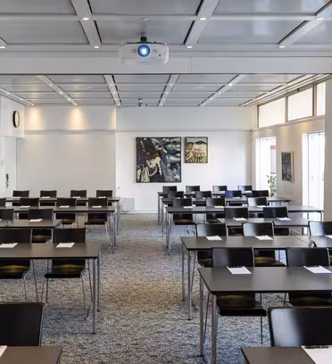 A conference room with rows of black chairs and tables arranged classroom-style. Each table has a notepad and pen. The room features a ceiling-mounted projector, two paintings on the back wall, and large windows with curtains on the side.