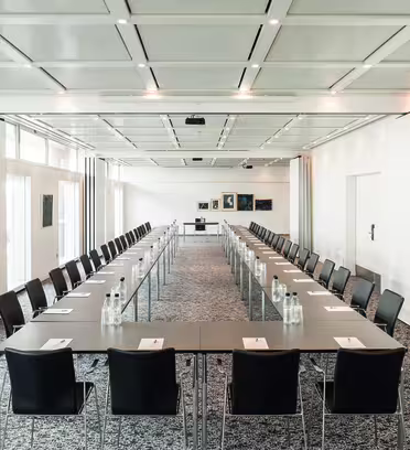 A large conference room with long tables arranged in a U-shape, lined with black chairs. Each table has a notepad and bottled water. The room has a patterned carpet, white walls, and several framed pictures at the far end. Ceiling lights are on.