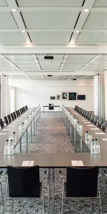 A large conference room with long tables arranged in a U-shape, lined with black chairs. Each table has a notepad and bottled water. The room has a patterned carpet, white walls, and several framed pictures at the far end. Ceiling lights are on.