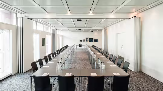 A large conference room with long tables arranged in a U-shape, lined with black chairs. Each table has a notepad and bottled water. The room has a patterned carpet, white walls, and several framed pictures at the far end. Ceiling lights are on.