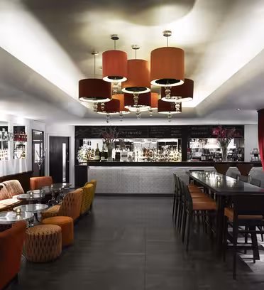 Modern bar interior with a sleek design featuring red-orange lighting fixtures. Seating includes orange and patterned chairs, high tables, and a long counter with black and white decor. Large black-and-white portraits adorn the walls.