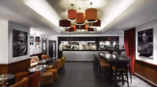 Modern bar interior with a sleek design featuring red-orange lighting fixtures. Seating includes orange and patterned chairs, high tables, and a long counter with black and white decor. Large black-and-white portraits adorn the walls.