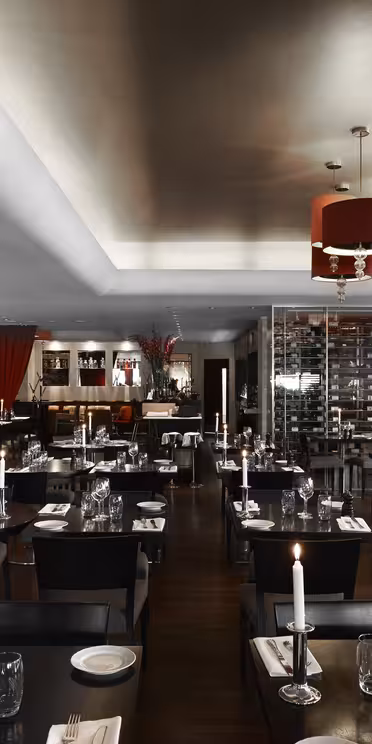 A modern restaurant interior with dark wooden tables set with glassware and napkins. Red cylindrical chandeliers hang from the ceiling. A wall features colorful murals, and a wine display is visible in the background.