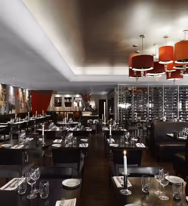 Elegant restaurant interior with dark tables set with candles and glassware. Large red lights hang from the ceiling. The walls feature modern artwork and a glass wine display is visible in the background.