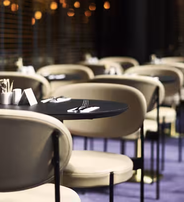 A modern restaurant with empty, elegantly set tables. Beige chairs line the black tables, each with cutlery and napkins. The floor is carpeted in purple, and the background features warm lighting and blinds.