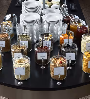 A buffet table with jars of cereals, nuts, and dried fruits, each labeled. Behind are stacks of white plates and cups, a basket of sugar packets, and plates of butter. Jars contain varied contents like granola and cornflakes.