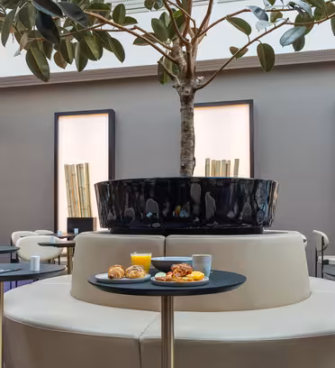 Modern dining area with light beige seating around a large potted tree. Tables hold breakfast items, including orange juice, pastries, and fruit. Tall, minimalist wall decor featuring bamboo accents in the background.
