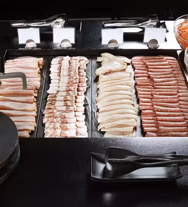 A buffet display featuring an assortment of sliced meats, including bacon, chicken, and sausage, arranged in rows. Nearby, bowls contain sliced tomatoes, cucumbers, and bell peppers. Serving tongs are placed in front.