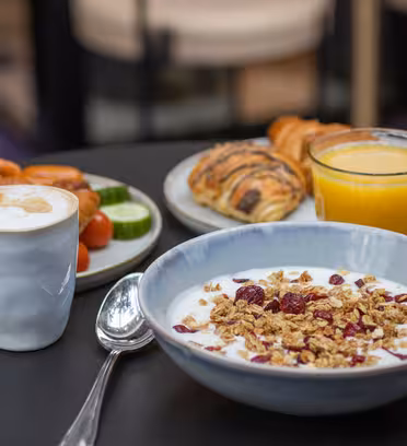 A breakfast spread on a table includes a bowl of yogurt with granola and berries, a cup of frothy cappuccino, a glass of orange juice, and a plate with a croissant, sausages, cheese, cucumber slices, and tomato.