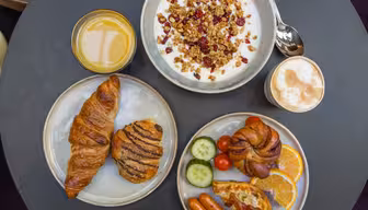 A round table with a breakfast spread: a bowl of yogurt topped with granola and berries, a glass of orange juice, a café latte, croissants, and a plate with tomatoes, cucumber slices, orange slices, sausages, bacon, and a bread roll.