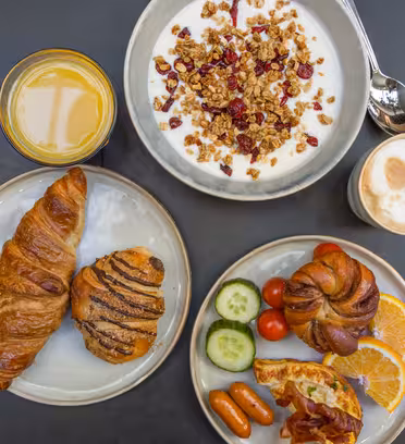 A round table with a breakfast spread: a bowl of yogurt topped with granola and berries, a glass of orange juice, a café latte, croissants, and a plate with tomatoes, cucumber slices, orange slices, sausages, bacon, and a bread roll.