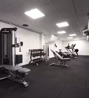 A modern gym featuring exercise machines, including a multi-station cable machine, a row of dumbbells on a rack, benches, and several stationary bikes and treadmills. The room has white walls, a black floor, and industrial ceiling ducts.