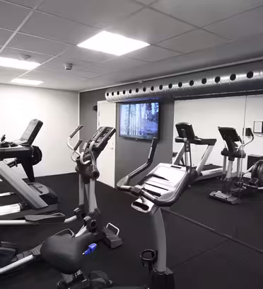 A modern gym room with treadmills, stationary bikes, and elliptical machines. The space has a mirrored wall, a ceiling with recessed lighting, and a metal ventilation duct. The floor is covered with black rubber mats.