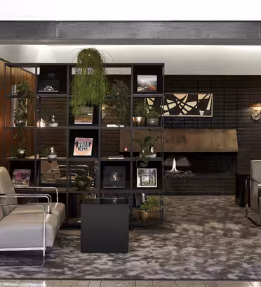 Modern lounge area with gray chairs and geometric shelves displaying plants and art. A unique fireplace is set against a dark brick wall. The space is accented with wood paneling and a patterned rug, creating a cozy, stylish atmosphere.