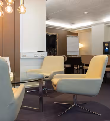Modern meeting room with beige chairs around a circular glass table. Warm ceiling lights hang above, and a flip chart and TV are visible in the background. The room has neutral tones and a carpeted floor.