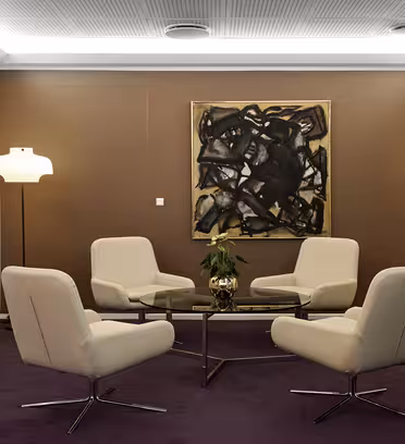A modern meeting room with four white swivel chairs surrounding a glass coffee table. Two floor lamps stand on either side of a large abstract painting on a brown wall. A flat-screen TV is mounted on the white wall. The carpet is a deep purple.