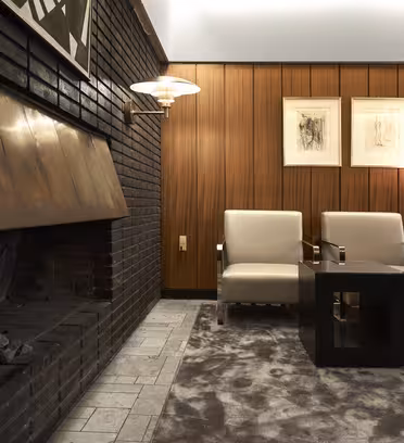 A cozy lounge with two beige armchairs and a square black table on a gray carpet. A lit fireplace with a copper hood is on the left, and two framed artworks hang on wood-paneled walls. A wall lamp and a pendant light illuminate the space.