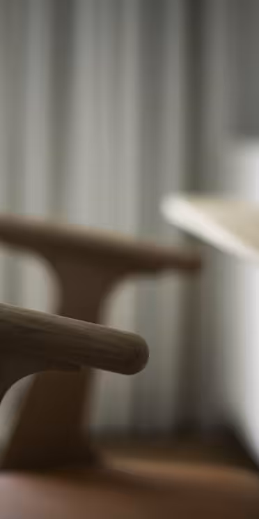 A wooden chair with a curved armrest is beside a wooden desk. In the background, a vase with green leaves is partially visible. The image is softly lit with natural light coming through a window with sheer curtains.